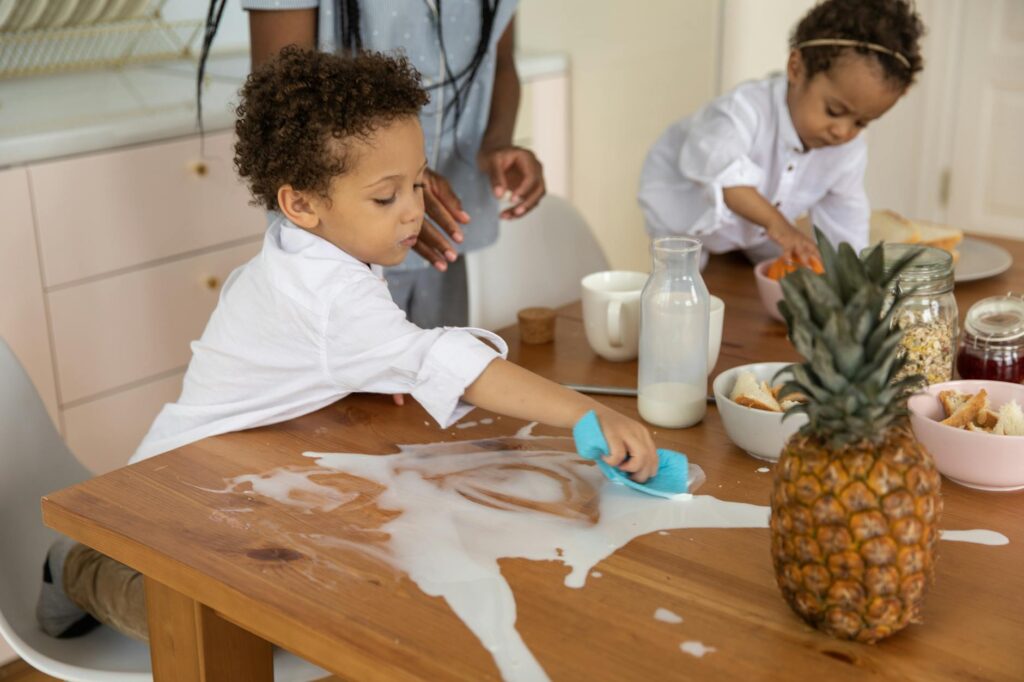 Children clean up a milk spill on a wooden breakfast table with family nearby, creating a cute and lively morning scene.